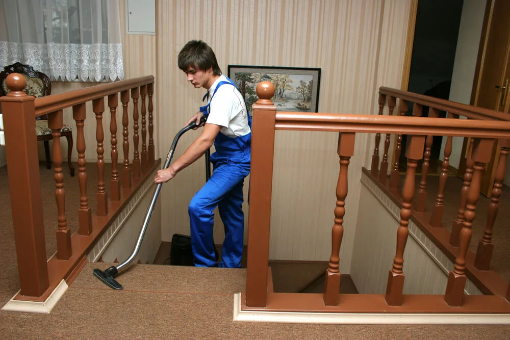 Cleaner performing carpet cleaning on stairs with a vacuum cleaner, wearing blue overalls and focusing on cleaning the carpeted surface in a home